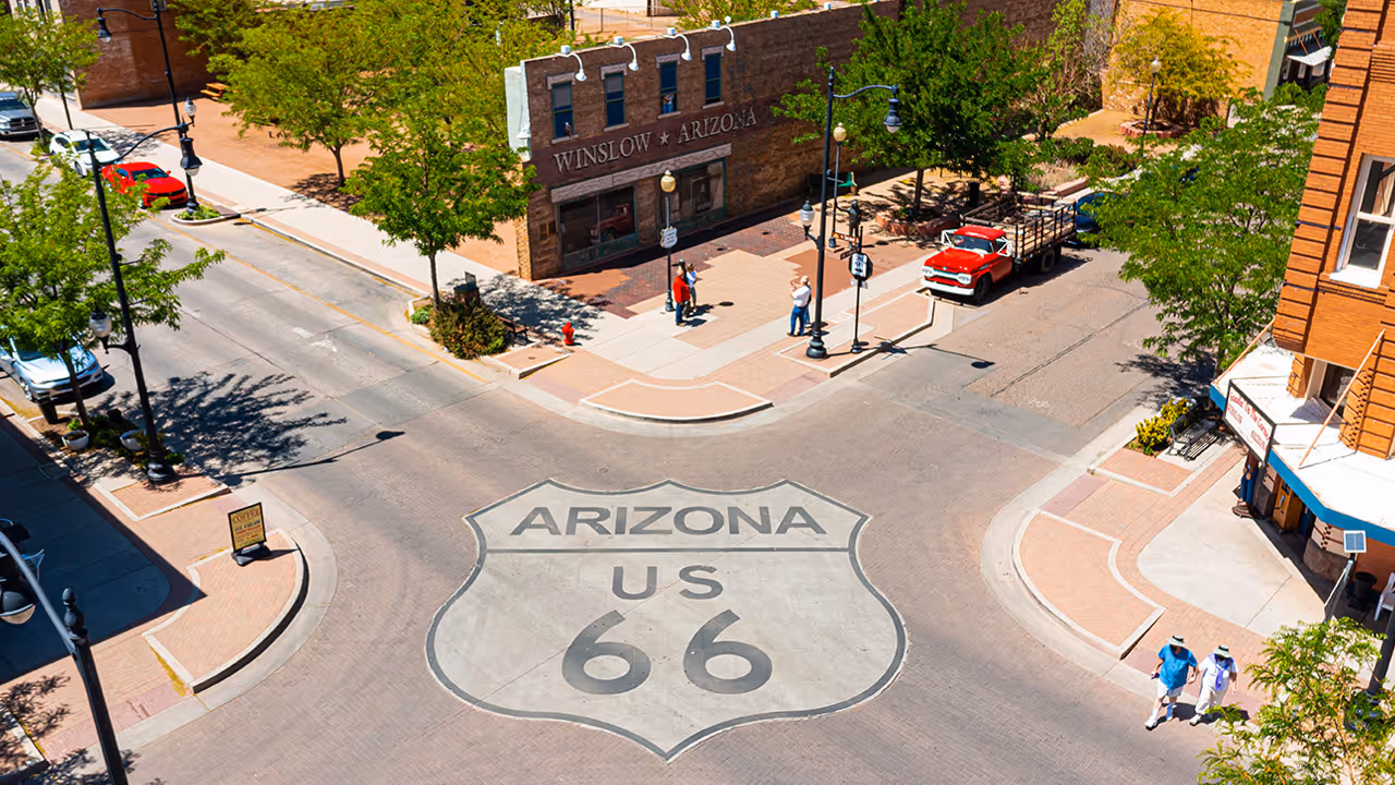 Aerial view of a street intersection in Winslow, Arizona, featuring a large Route 66 shield painted on the road with a vintage red pickup truck and people walking nearby.